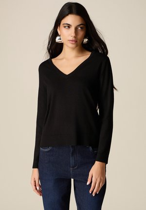 Kobieta w czarnym swetrze z długim rękawem i dekoltem w kształcie litery V, niebieskich dżinsach oraz dużych srebrnych kolczykach kołach na jasnym, jednolitym tle.