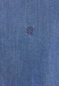 Tessuto di denim in una tonalità blu medio con un piccolo logo geometrico ricamato in un filo più chiaro sul lato sinistro inferiore.