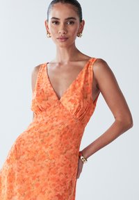 Oranžna cvetna obleka z V-izrezom, poudarjenim pasom in lahkotno krilom. Izdelana iz lahkega blaga z mehko teksturo. Zlati obroči za ušesa.