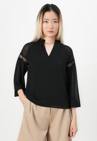 Blusa nera con scollatura a V, dettagli arricciati e maniche trasparenti con inserti in pizzo. Il tessuto ha una texture leggera e liscia.