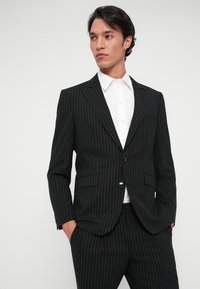 Μαύρο σακάκι με ραβδώσεις και κοπή που ταιριάζει. Διαθέτει μπροστινό κλείσιμο με ένα κουμπί, πέτα με εγκοπή και δύο μπροστινές τσέπες, συνδυασμένο με λευκό πουκάμισο.