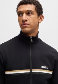 Schwarze Zip-Jacke mit horizontalen Streifen in Gold, Weiß und Schwarz. Der Markenname "BOSS" ist in Weiß auf der Brust aufgedruckt.
