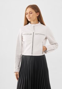 Vit läderjacka med hög krage, dragkedja fram och fyra dragkedjefickor. Modellen bär en veckad svart kjol.