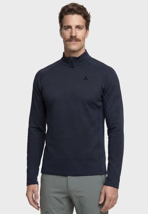 Mann mit braunen Haaren und Schnurrbart, trägt ein marineblaues Langarmoberteil mit Viertelreißverschluss und hellgraue Hose, steht vor einem einfarbigen Hintergrund.