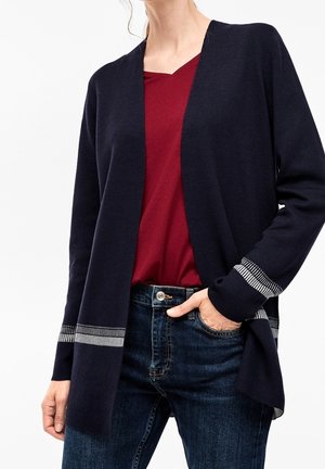 Cardigan lungo navy con apertura frontale, polsini a coste con strisce grigie, indossato su una maglietta borgogna e jeans scuri. Tessuto morbido e leggero.