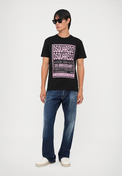 Camiseta negra con texto gráfico rosa, "DSQUARED2 30TH ANNIVERSARY", combinada con jeans de mezclilla azules y zapatillas blancas. Modelo con gafas de sol negras.