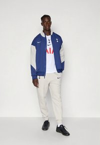 Navy and light gray jacket with a zip, white Spurs logo shirt, beige joggers, and black shoes. Minimalist sportswear design.