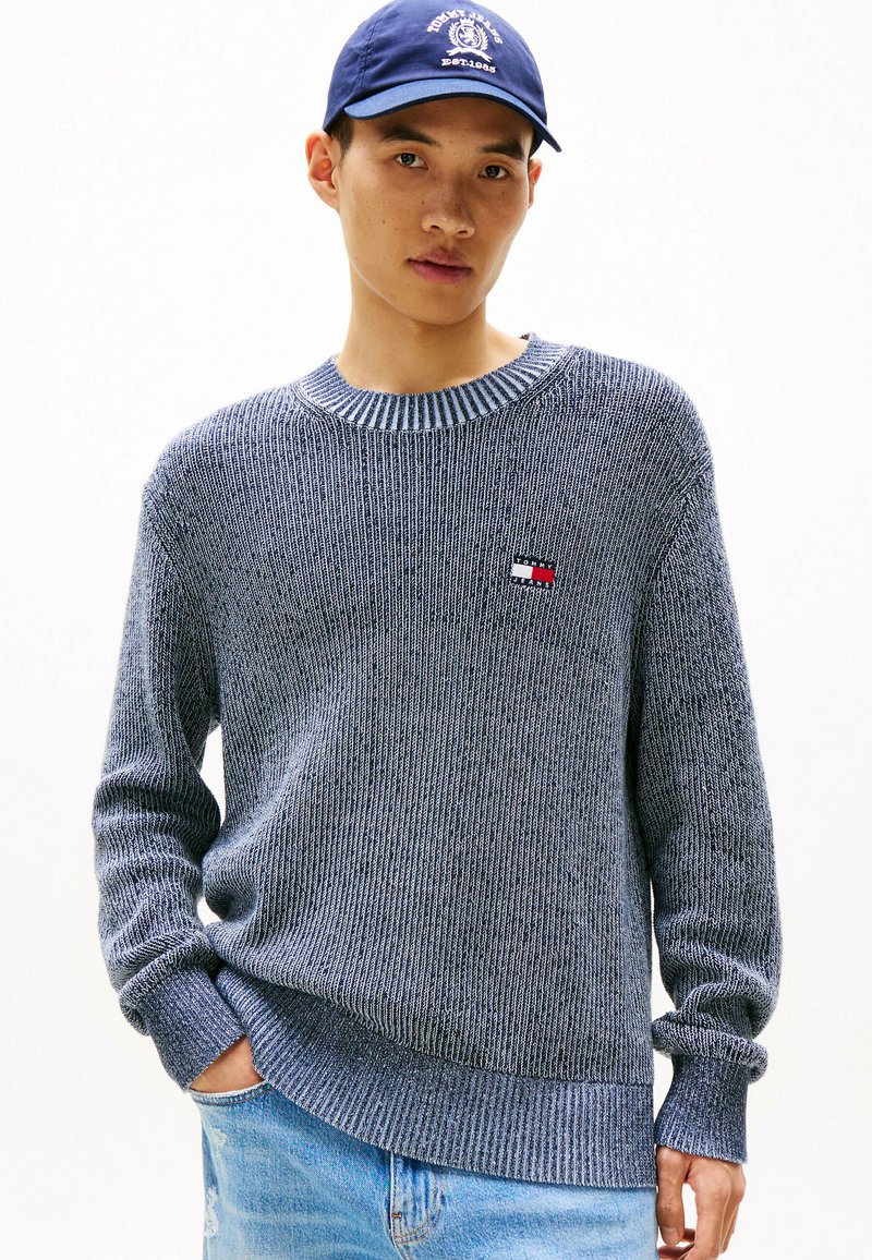 Grauer gerippter Strickpullover mit Rundhalsausschnitt und langen Ärmeln. Verfügt über ein kleines Logo-Patch auf der Brust und hat einen locker sitzenden Schnitt. Trägt dazu eine hellblaue Jeans.
