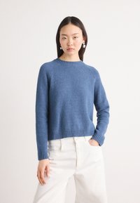 Blauwe gebreide trui met een geribbelde ronde hals en manchetten, met een relaxte pasvorm. Gecombineerd met witte hoge taille broeken.