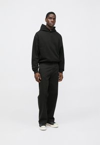 Čierna mikina s kapucňou s predným vreckom na puky a šnúrkou na stiahnutie, v kombinácii s čiernymi širokými nohavicami a čiernymi teniskami s bielymi podrážkami.