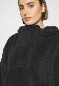 Schwarzer Fleece-Hoodie mit hohem Kragen, weichem Material und einer aufgesetzten Tasche auf der Vorderseite. Gemütliches und oversized Design.
