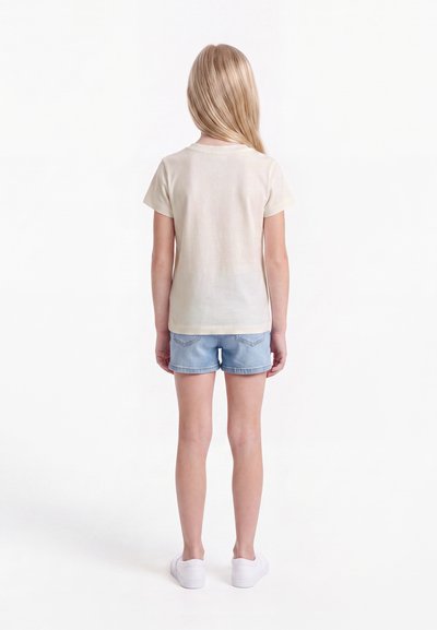 Enfant aux longs cheveux blonds debout de dos portant un t-shirt beige, un short en jean bleu clair et des baskets blanches sur fond blanc.