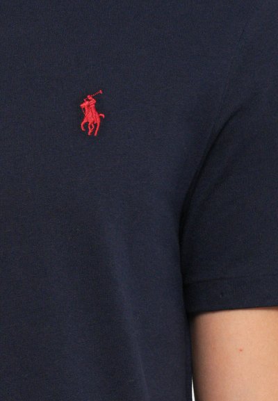 Mornarsko modro blago s rdečim vezenim logotipom polo igralca na konju, posnetek v bližini zgornjega dela prsnega koša in rokava kratkosleče majice.