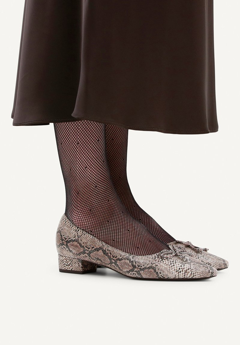 Chaussures plates avec un motif python, dotées d'un bout arrondi et d'un détail nœud. Portées avec des collants noirs en résille et une jupe marron.