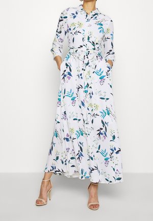 Kobieta ubrana w długą białą sukienkę z niebieskimi, zielonymi i fioletowymi kwiatowymi nadrukami, zapinaną na guziki z przodu, z paskiem w talii i beżowymi sandałami na wysokim obcasie.