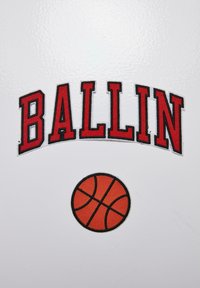 Raudona siuvinėta tekstas "BALLIN" išlenktas virš oranžinio krepšinio pačios su juodomis linijomis, esančiame baltame fone.