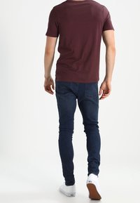 Man som går bort iförd en burgunderröd kortärmad skjorta, mörkblå slim-fit jeans och vita sneakers mot en vit bakgrund.