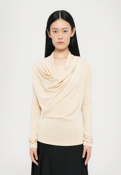 Beige long-sleeve top with a draped cowl neckline. Smooth, stretchy fabric; fitted design enhances shape. Worn with a black skirt.
