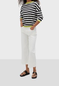 Gestreiftes Strickoberteil in Schwarz-Weiß mit einem Limettengrünen Saum, kombiniert mit weiten weißen Hosen und schwarzen Sandalen mit goldenen Akzenten.