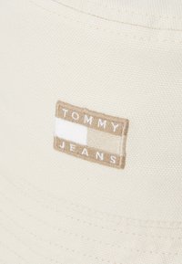 Kremasti šešir s uskim rubom izrađen od teksturirane tkanine. Sadrži pravokutnu bež oznaku s natpisom "TOMMY JEANS", ukrašenu bijelom prugom.