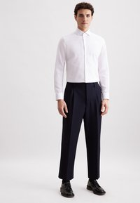 Uomo in camicia bianca con bottoni, pantaloni neri a pieghe, calze nere e scarpe eleganti nere, in piedi davanti a uno sfondo semplice.