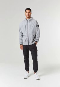 Jasnoszara kurtka z kapturem z dwoma przednimi kieszeniami, noszona na czarnych spodniach cargo i białych trampkach, z naszywką logo na rękawie.