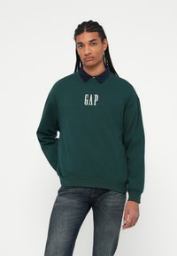 Felpa verde con logo "GAP" bianco, caratterizzata da un colletto blu scuro. Indossata con jeans scuri, mostra una vestibilità rilassata e una morbida consistenza.