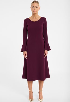 Frau in einem langärmeligen, dunkelvioletten Midikleid mit ausgestellten Bündchen, die aufrecht steht, kombiniert mit metallisch goldenen Riemchen-Heels.