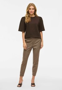 Brūns īsās piedurknes tops ar savilktiem pleciem, apvienots ar pieguļošām brūnām biksēm. Modelis valkā tumšas papēžu sandalītes. Vienkāršs, moderns dizains.