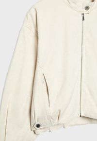 Hellbeige Jacke mit Frontreißverschluss, seitlichen Taschen, Druckknopfdetail am Saum und langen Ärmeln vor einfarbigem Hintergrund.