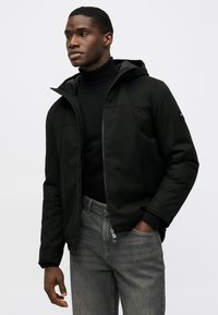 Čierna kapucňová bunda na zips, s hladkým povrchom, bočnými vreckami a ribovanými manžetami; skombinovaná so šedými džínsami.