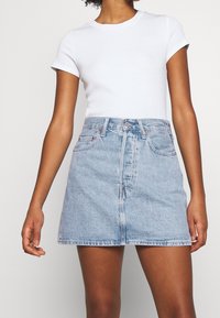 Femme portant un t-shirt blanc uni à manches courtes et une mini-jupe en denim bleu clair taille haute avec poches avant et fermeture par boutons.