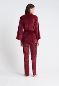 Robe en velours bordeaux avec ceinture à nouer, manches longues et pantalon assorti à motif floral dans une teinte similaire. Texture douce, coupe décontractée.
