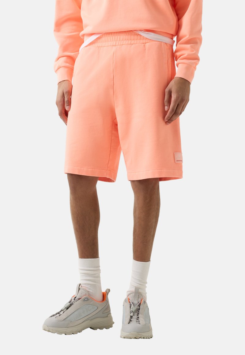 Mand iført ferskenfarvede shorts, matchende sweatshirt, hvide sokker og grå sneakers, stående mod hvid baggrund.