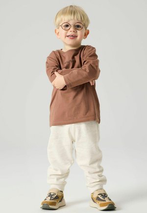 Young boy with blonde hair and glasses, wearing a brown long-sleeve shirt, white pants, and yellow sneakers, standing with arms crossed and smiling.