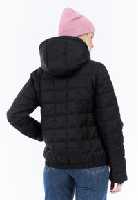 Donna che indossa una giacca nera trapuntata con cappuccio, jeans blu e un berretto di maglia rosa, in piedi contro uno sfondo bianco.