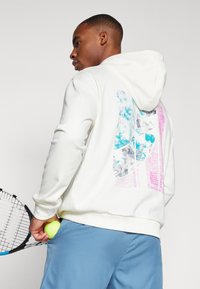 Hvit hettegenser med et fargerikt grafisk motiv som viser blå, rosa og grå teksturer på baksiden. Modellen holder en tennisrakett og en ball.