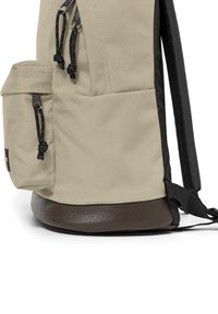 Béžový plátěný batoh s černým lemem, texturovaným hnědým dnem a boční kapsou s černými stahovacími detaily a kovovými akcenty.