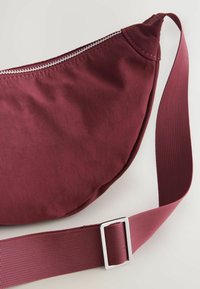 Burgundy crossbody taske i blødt, struktureret stof med lynlås og justerbar rem, prydet med en metal spænde accenter.