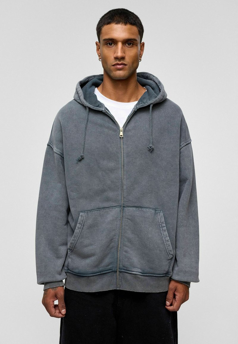 Grijze zip-up hoodie met een gestructureerde afwerking, grote voorkruippockets, verstelbare trekkoordcapuchon en ribbels bij de mouwen. Eenvoudig ontwerp met een ontspannen pasvorm.