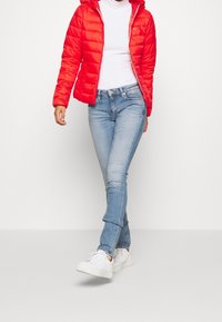 Persona che indossa un giubbotto imbottito rosso, un dolcevita bianco, jeans azzurri chiari e sneakers bianche, con una gamba incrociata davanti.