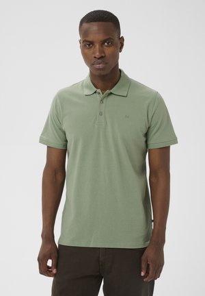 Egy férfi világoszöld rövid ujjú galléros pólót és sötét nadrágot visel, egy egyszerű világos háttér előtt áll.