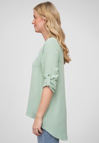 Top verde menta a maniche lunghe con vestibilità rilassata, tessuto testurizzato, polsini arrotolati e orlo leggermente curvo. Abbinato a jeans blu.