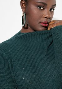 Mörkgrön ribbade tröja med hög hals och metalliska inslag. Modellen bär stora hoopörhängen och lätt definierade läppar.