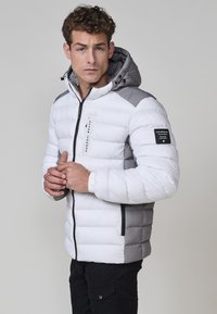 Giacca imbottita bianca e grigia, dotata di cappuccio, chiusura con zip e polsini elastici. Dettaglio del logo sulla manica. Texture liscia e design cucito.