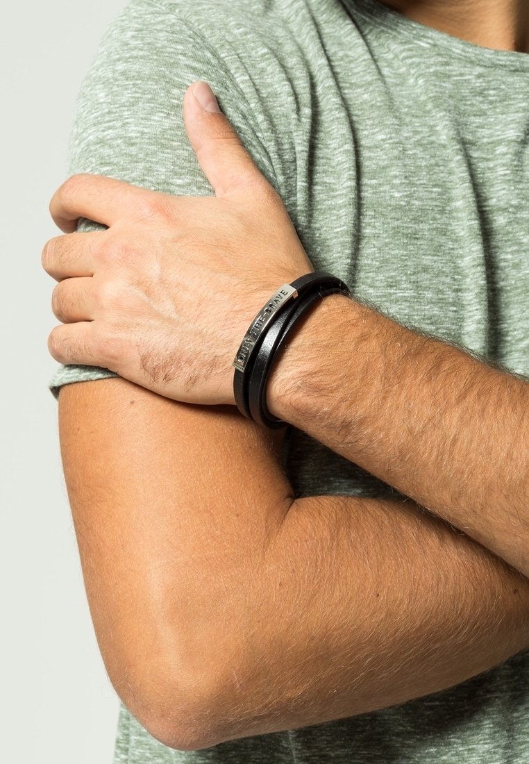 Bras croisés d'un homme portant une chemise à manches courtes vert clair chinée et un bracelet en cuir noir avec une plaque métallique portant l'inscription "DON'T LET GRAVE".