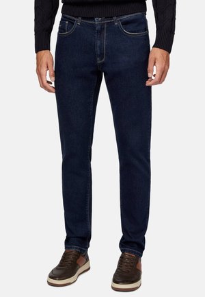 Mann trägt dunkelblaue Slim-Fit-Jeans kombiniert mit braunen und beigen Sneakern und einem schwarzen Pullover.