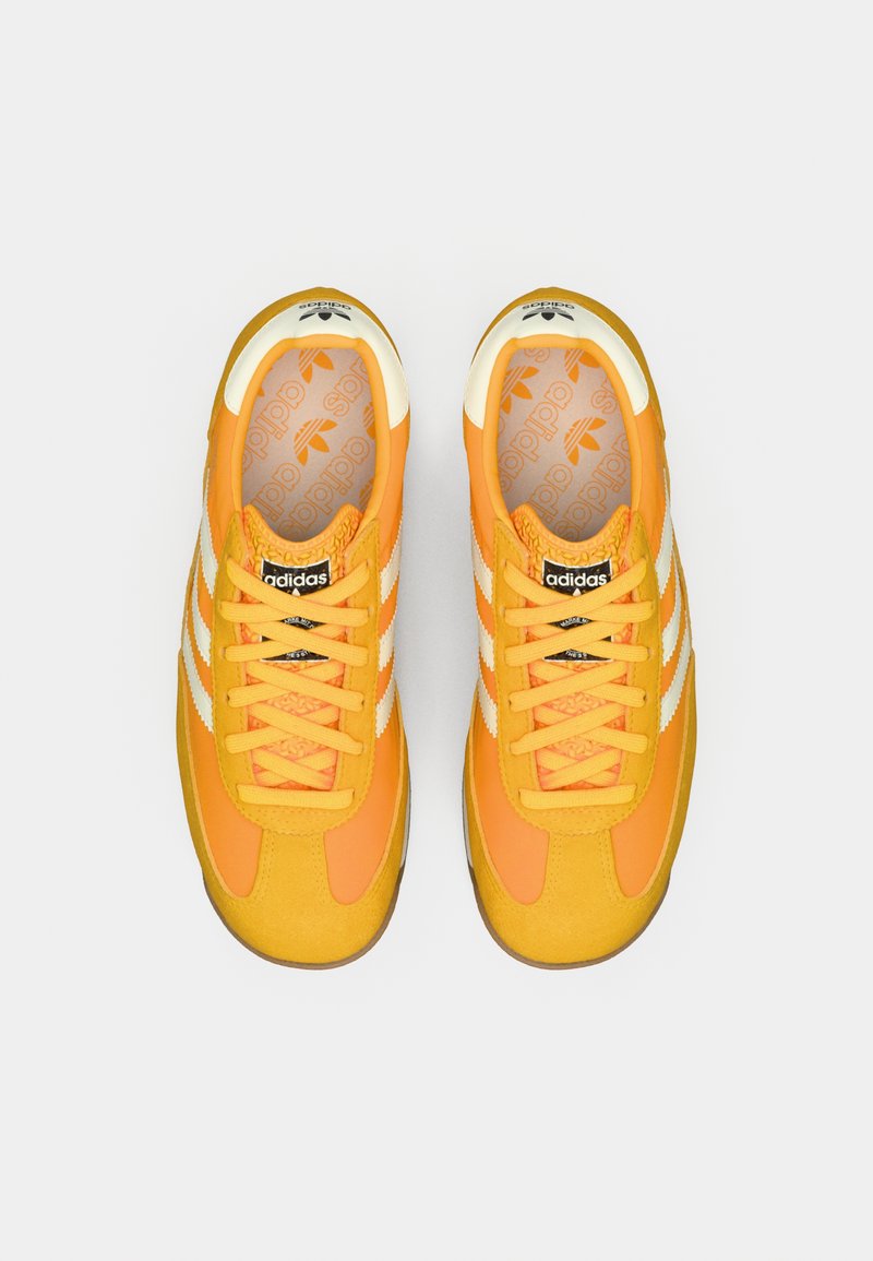 Un par de zapatillas Adidas de un brillante color naranja con rayas y cordones blancos, vistas desde arriba sobre un fondo gris liso.