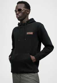 Zwarte hoodie met een voorzak, een trekkoordcapuchon en een kleurrijk "RIPCURL" logo op de borst. Glad materiaal, strak ontwerp, gedragen met lichte broek.