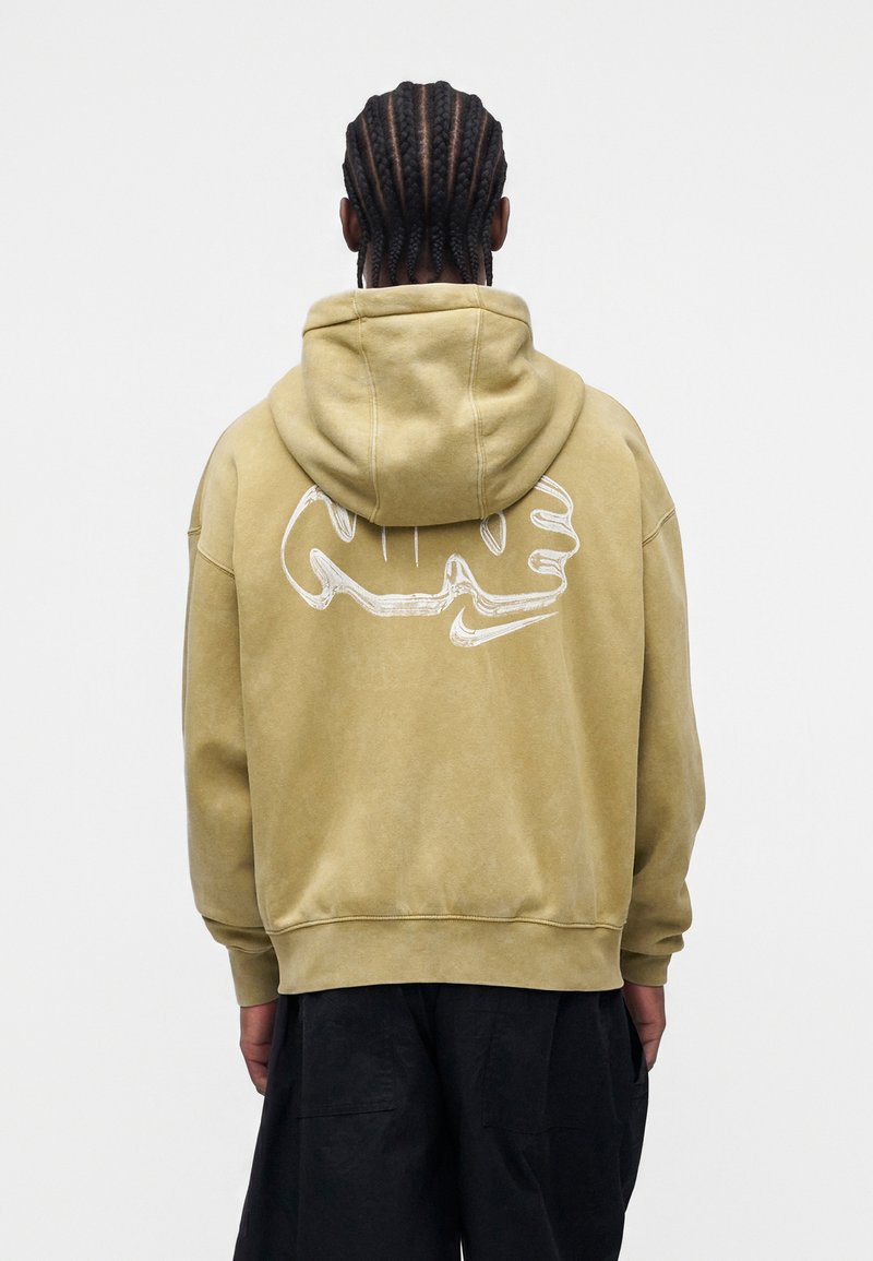 Person mit geflochtenem Haar, die einen beige Hoodie mit weißem abstraktem Gesicht und Nike-Swoosh-Design auf dem Rücken trägt, kombiniert mit dunkler Hose.